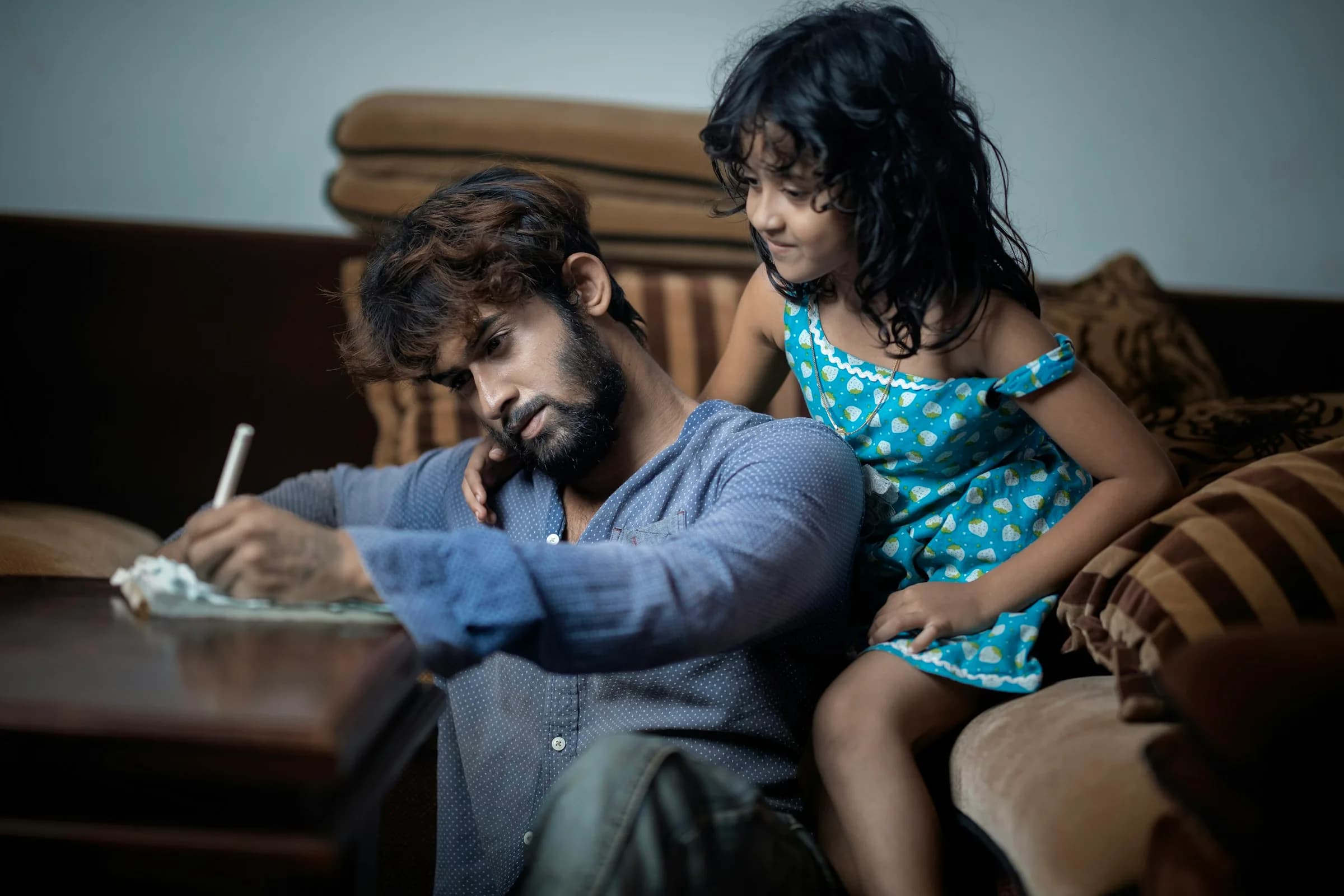 Padre llenando documentos en la mesa de la sala mientras su hija pequeña lo observa